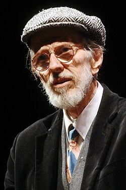 An elderly man with a white beard, round glasses, xand flat cap.
